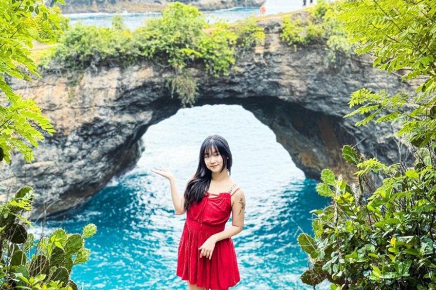 A person in a red dress standing in front of a natural rock arch over water. 2