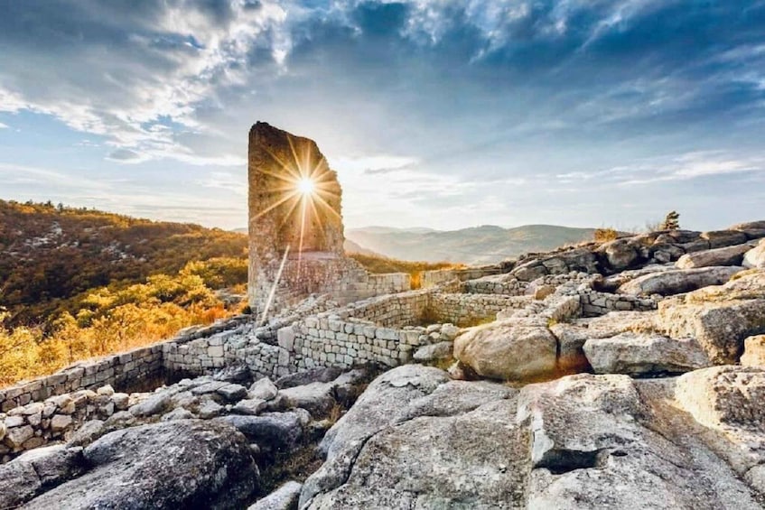 A stone ruin with a bright sun shining through a gap in the structure, surrounded by rocky terrain and autumn-colored trees. 1