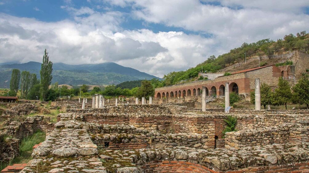 Ancient ruins with stone walls and columns, set against a backdrop of mountains and a cloudy sky. 1