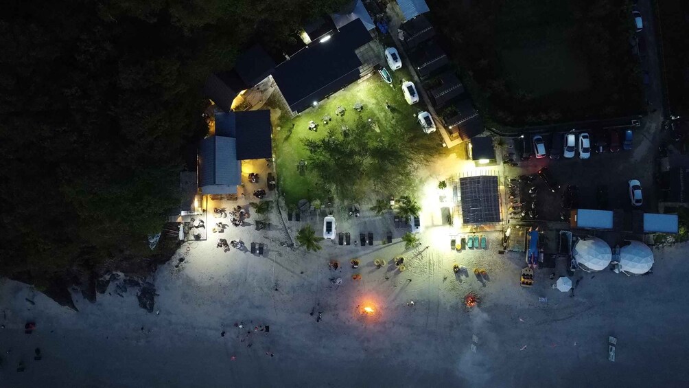 A nighttime scene with a lit building, parked cars, and a sandy area. 2
