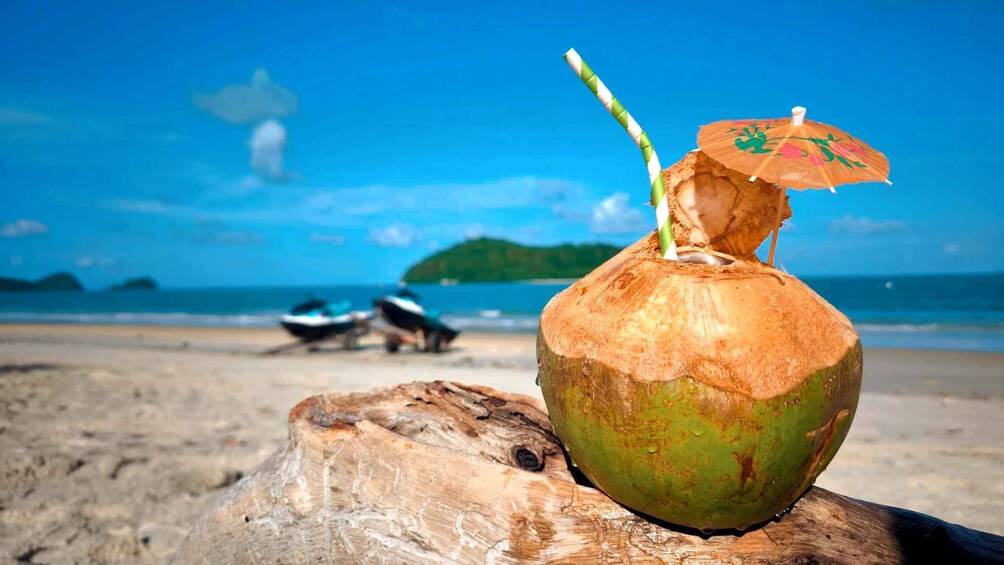 A coconut on a wooden stump with a straw and a colorful umbrella on top, set against a beach with boats and a clear blue sky. 5
