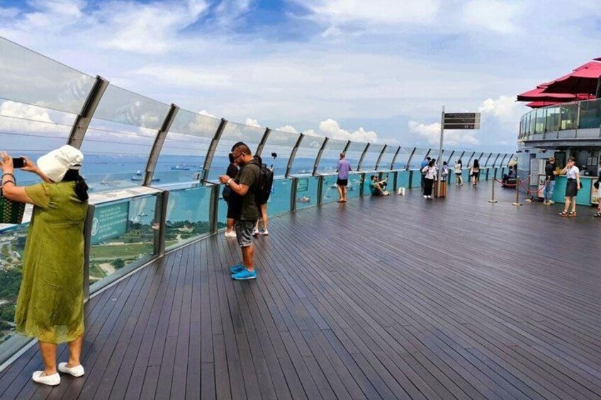 A deck with glass railings overlooking the ocean, people taking photos, and a ship in the background. 5
