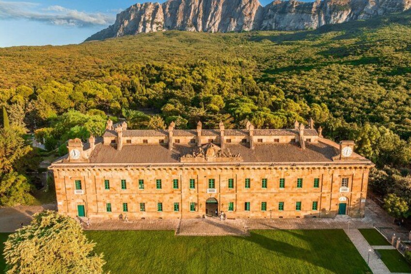 A large, historic building with a symmetrical facade, surrounded by lush greenery and mountains in the background. 3