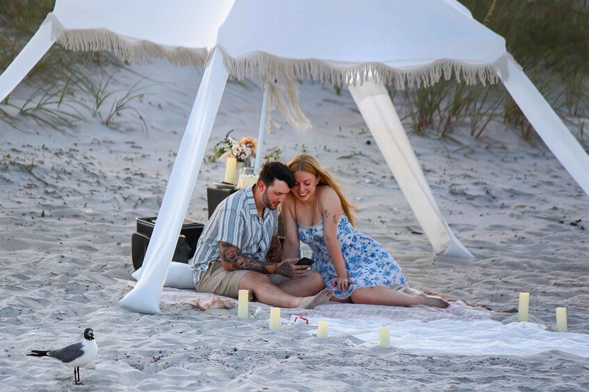 A couple sitting under a white tent on a sandy beach with candles and a bird nearby. 5