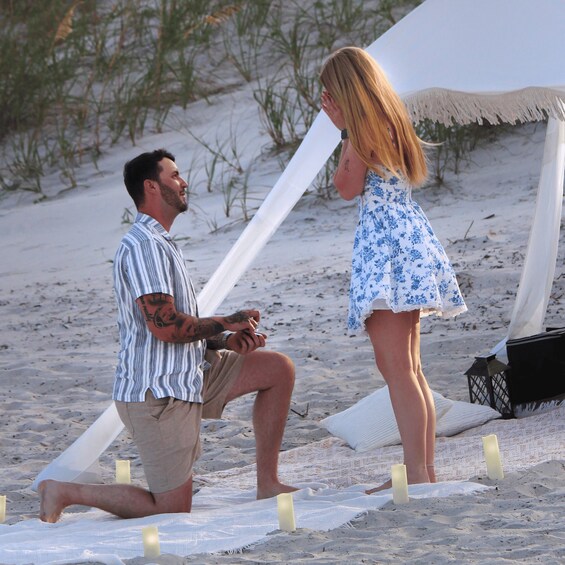 A man kneeling on a beach with a ring, proposing to a woman standing nearby. 2