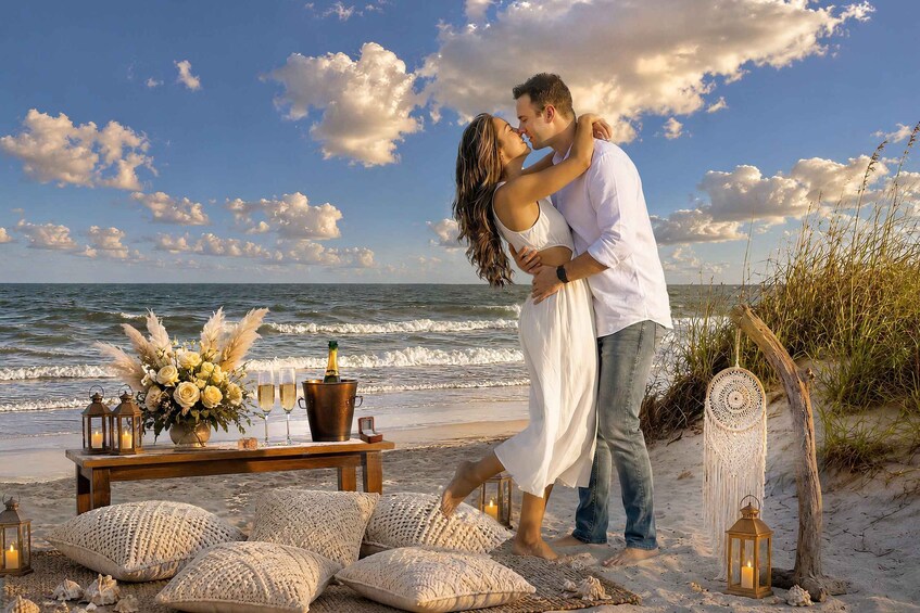 A couple embracing on a beach with a table of drinks, flowers, and lanterns. 1
