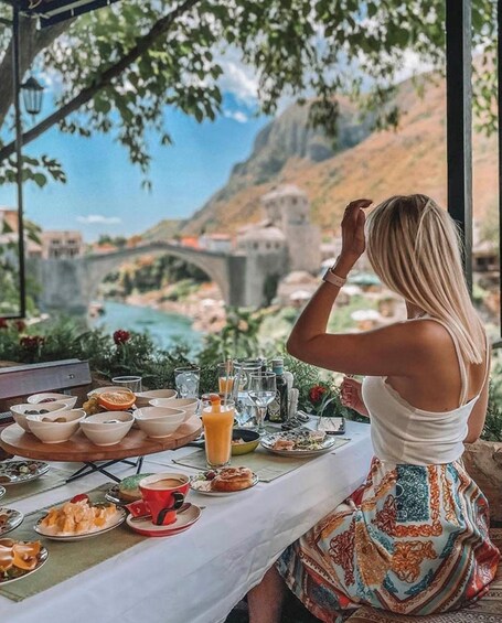 A woman at a table with a meal, overlooking a scenic view of a bridge and mountains. 1