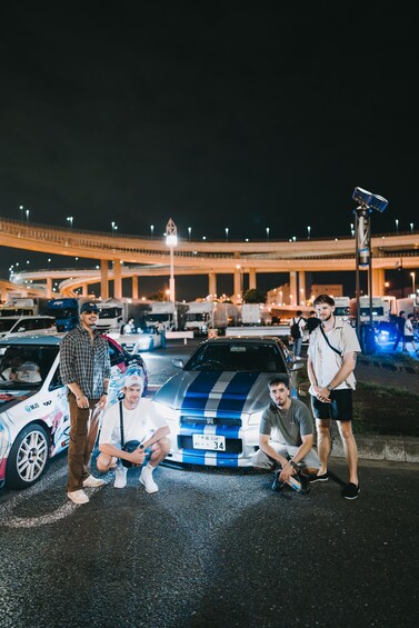 Een groep mensen poseert voor een blauw-wit gestreepte auto in de nacht. 2