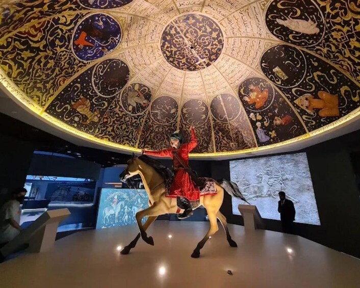 A museum exhibit with a person in a red outfit riding a horse, surrounded by a large, ornate ceiling with circular designs and inscriptions. 5