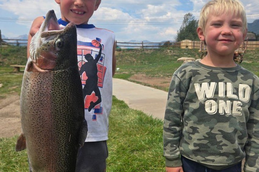 Two children holding a large fish, standing on a grassy area with a fence and trees in the background. 2