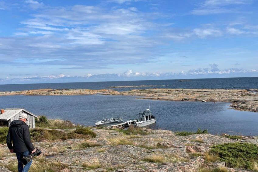 A rocky shoreline with a boat docked on a calm body of water, a person standing on the shore, and a distant mountain range under a partly cloudy sky. 5
