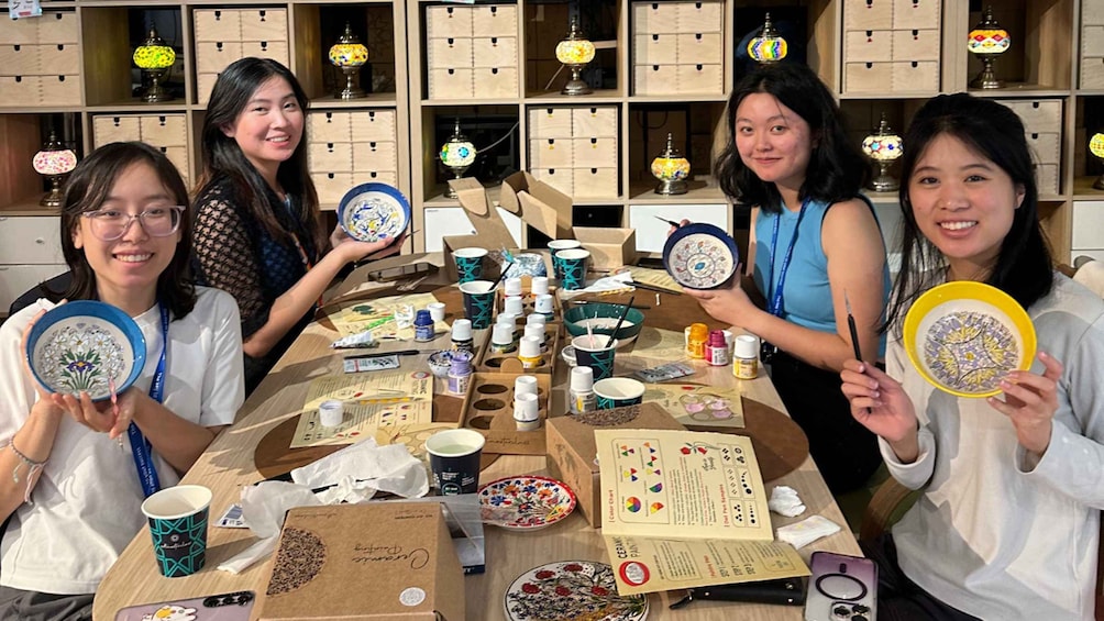 A group of people at a table with pottery and paint supplies, surrounded by shelves with decorative lamps. 1