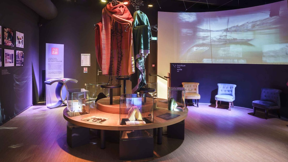A museum exhibit with a central display of textiles and a projection screen in the background. 2