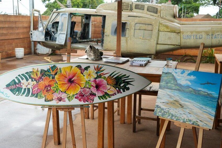 A table with a floral painting, an easel with a landscape painting, and a vintage military vehicle in the background. 3