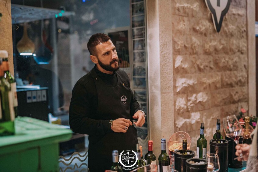 A person in a black uniform with a logo, standing in a bar with various bottles and glasses on the counter. 3