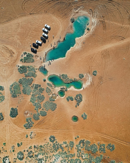 An aerial view of a desert landscape with a series of turquoise pools, scattered vegetation, and a row of vehicles parked near the pools. 1