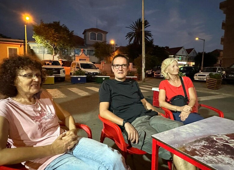 Tre personer sitter vid ett bord utomhus i skymningen, med gatlyktor och byggnader i bakgrunden. 5