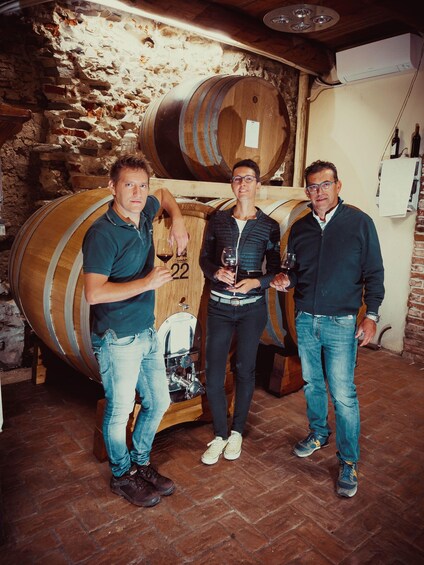 Three people standing in a cellar with wooden barrels, holding glasses of wine. 4