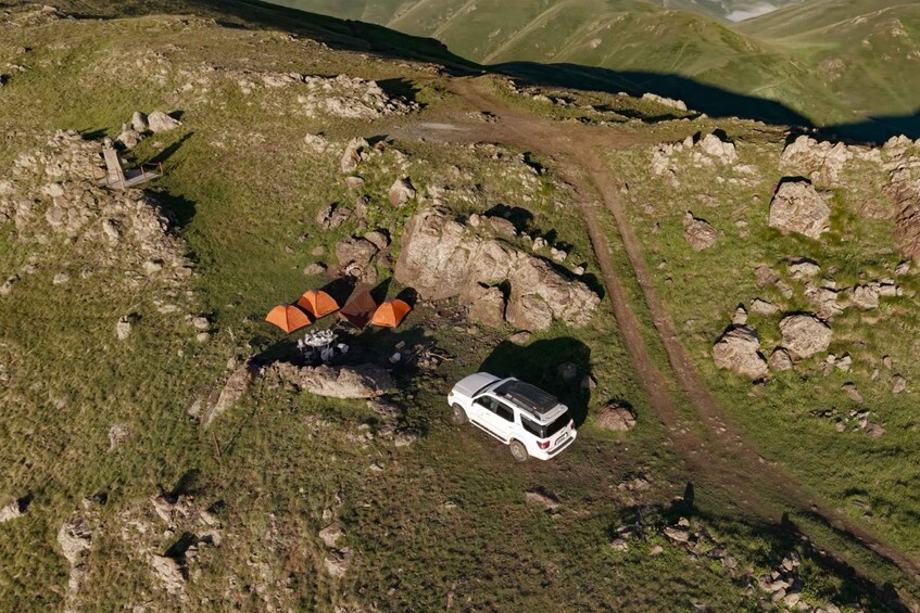 Eine Luftaufnahme eines gebirgigen Gebiets mit einem weißen Fahrzeug, das in der Nähe einer Felsformation geparkt ist, umgeben von Gras und einem Feldweg. 5