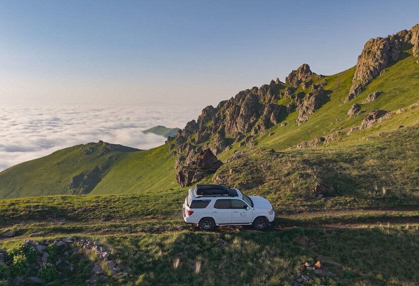 A white SUV parked on a grassy hillside with rocky terrain and a view of clouds below. 1