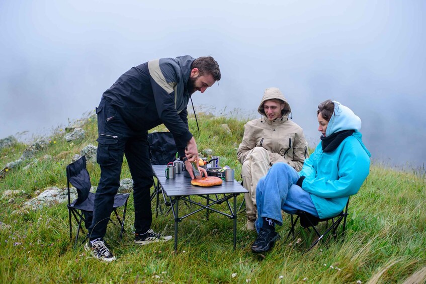 Three people outdoors, one preparing food on a portable stove, with a grassy field and foggy sky in the background. 4