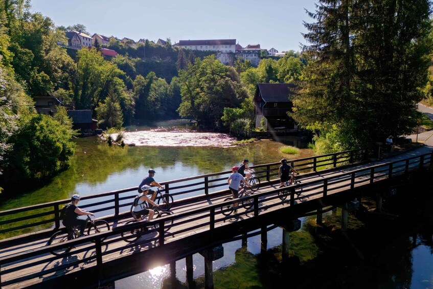 A group of people cycling on a wooden bridge over a river, surrounded by greenery and buildings in the background. 2
