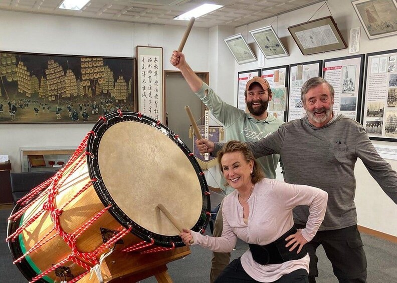 Three people in a room with a large drum, framed pictures, and a map on the wall. 1