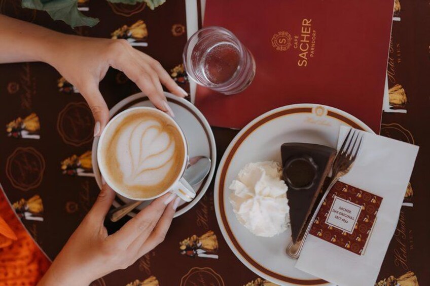 A person holding a cup of coffee with a heart-shaped latte art, a plate with a chocolate dessert, and a menu on a table with a patterned tablecloth. 2