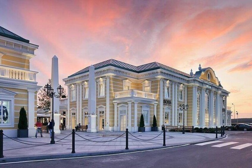 A classical building with columns and a large central entrance, surrounded by a paved area and a street. 1