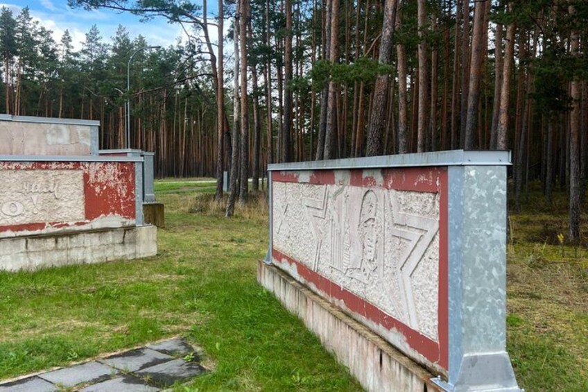Two weathered memorial plaques with inscriptions, set in a grassy area with a forest in the background. 2