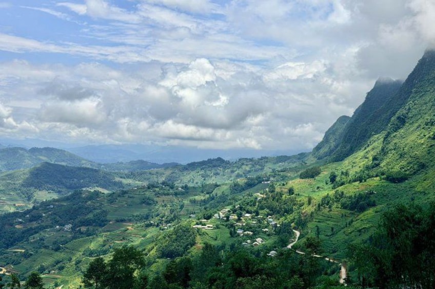A lush green valley with terraced fields, a winding road, and distant mountains under a partly cloudy sky. 1