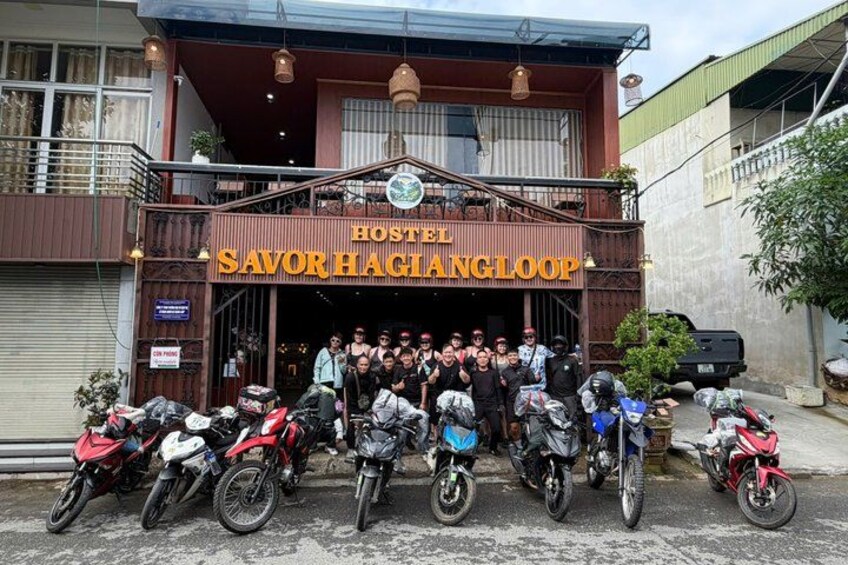 A group of people posing in front of a building labeled 'HOSTEL SAVOR HAGIANGLOOP' with several motorcycles parked in front. 3