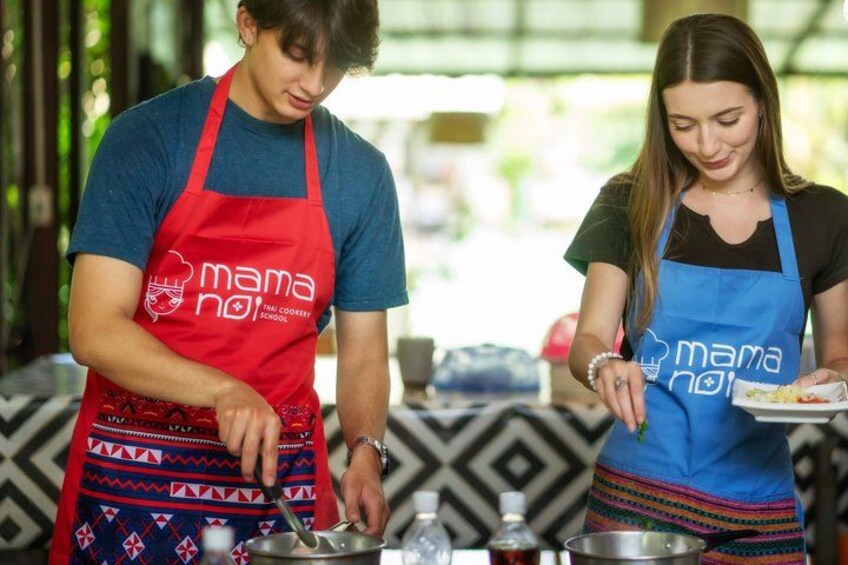 Two people wearing aprons with 'mana no' logos, cooking outdoors. 1