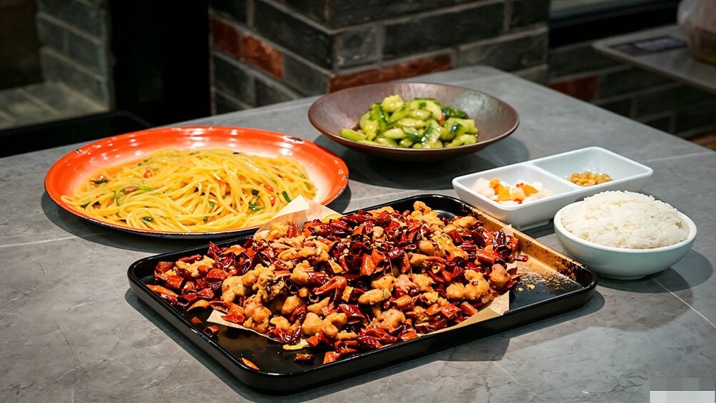 Ein Tisch mit einer Auswahl an Speisen, darunter eine Portion Spaghetti, eine Platte mit gebratenem Fleisch und Paprika, eine Schüssel mit Gurkenscheiben sowie kleine Schälchen mit Reis und weiteren Beilagen. 4