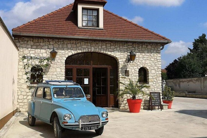 A vintage blue car parked in front of a stone building with a red roof, potted plants, and a signboard. 3