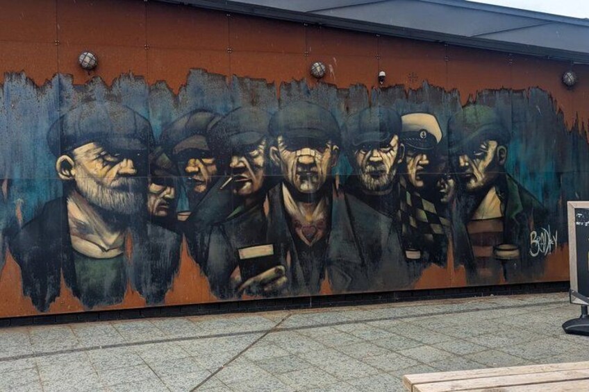 A mural with multiple figures, a bench, and a trash bin. 4