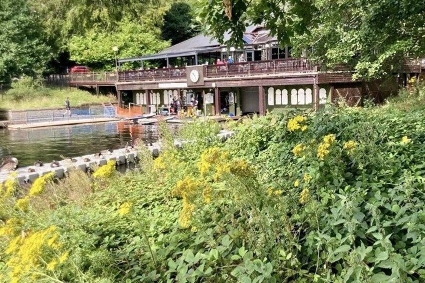 A riverside building with a balcony overlooking the water, surrounded by greenery and yellow flowers. 2