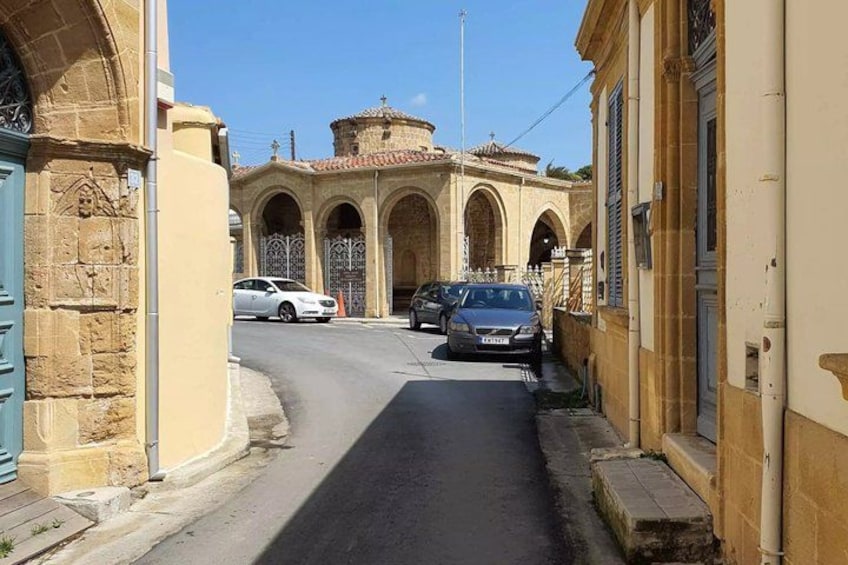 A narrow street with parked cars, a building with arched doorways, and a tower in the background. 4