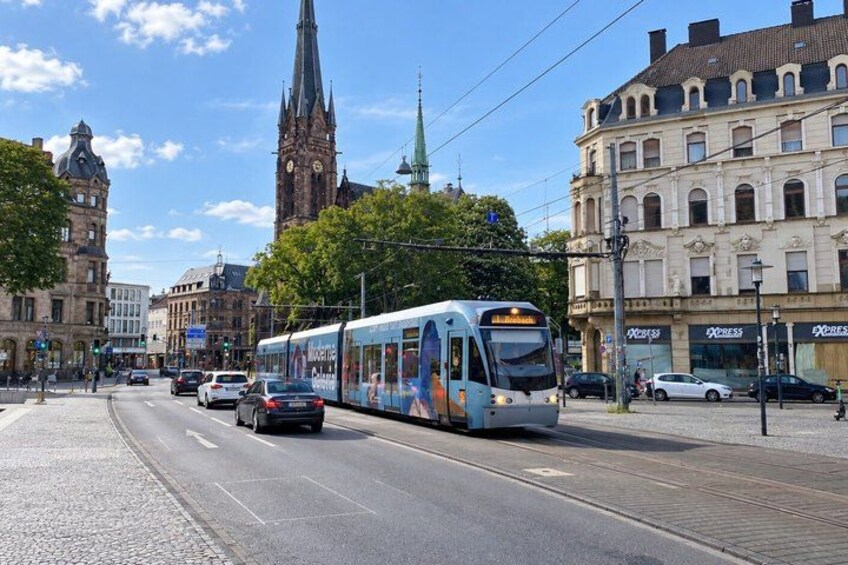 A tram on a city street with buildings and a church tower in the background. 2
