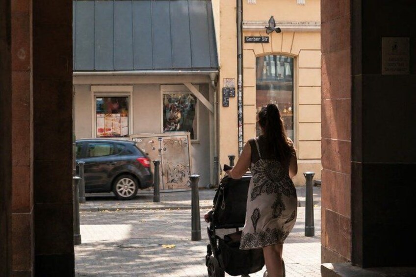 A woman walking with a stroller on a sidewalk, a parked car, and a building with a sign that reads 'Serber St.' 5