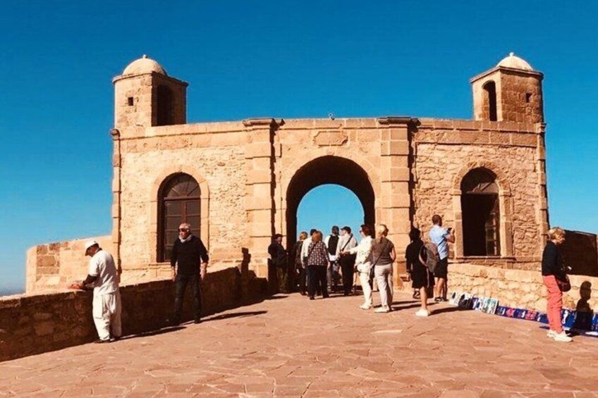 A stone archway with tourists, a clear sky, and a building with a tower. 1