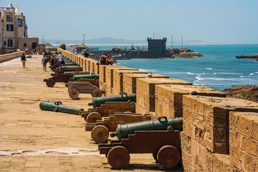 A coastal promenade with cannons lined up along the wall, a clear sky, and a distant view of the sea. 2