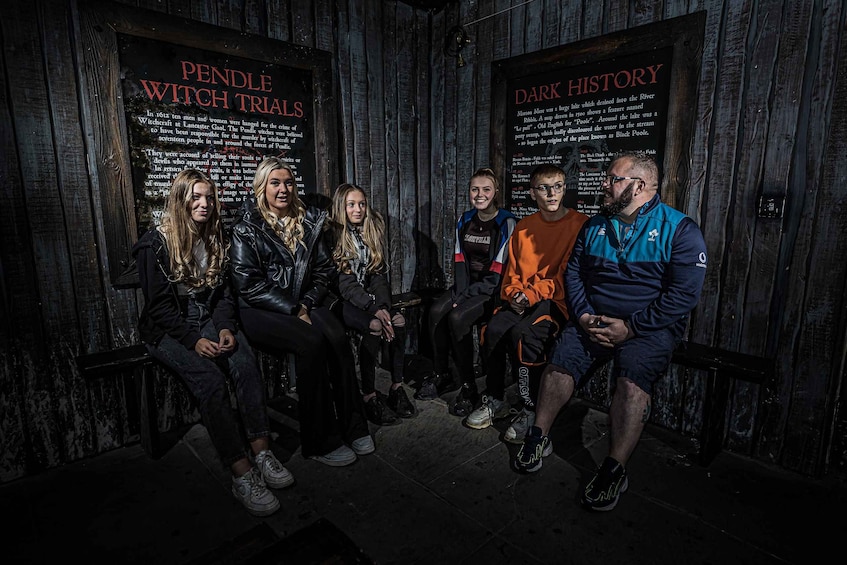 A group of people sitting on benches in front of informational signs about Pendle Witch Trials and Dark History. 3