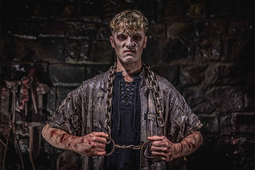 A person in a dark, chain-bound outfit with a rustic background. 4