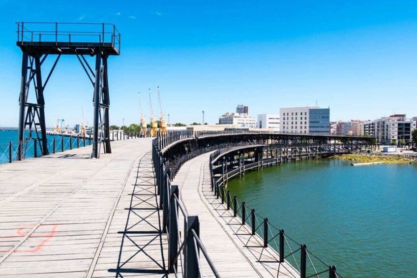 A waterfront promenade with a curved walkway, a metal structure on the left, and a city skyline in the background. 1