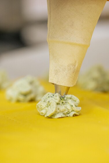 A hand using a piping bag to apply a creamy, herb-infused substance onto a yellow surface. 5