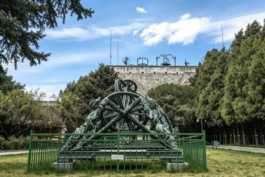 A large metal sculpture in the shape of a wheel with a central figure, surrounded by a green fence, in a park with trees and a building in the background. 1