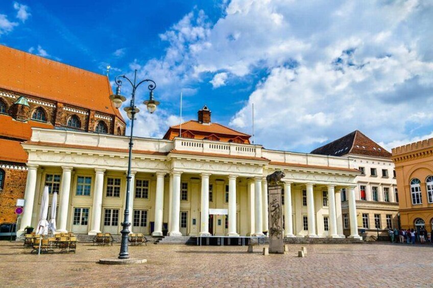A historic building with classical architecture, a cobblestone plaza, and a clear blue sky. 3