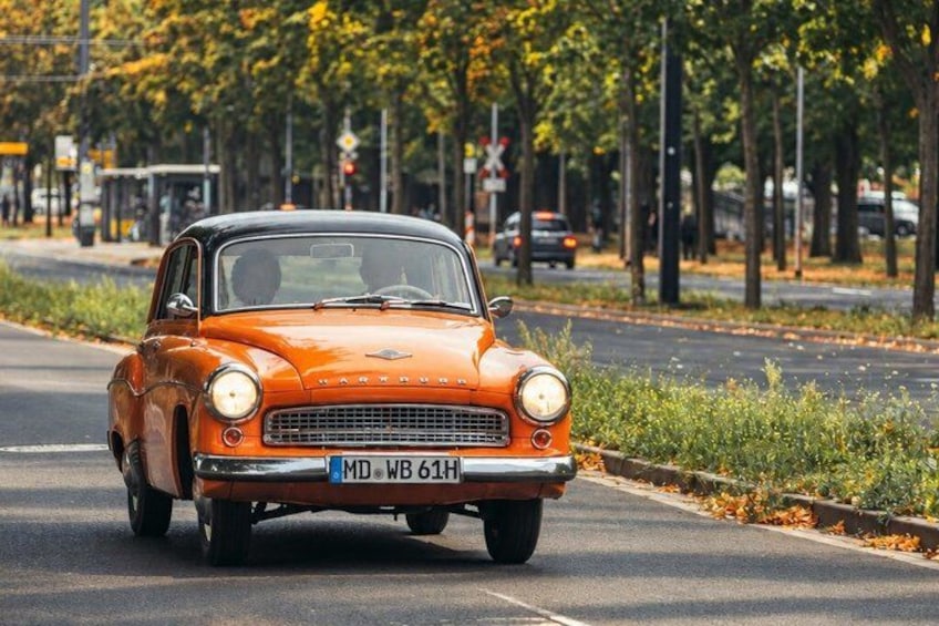 An orange vintage car driving on a road with trees and a pedestrian walkway. 4