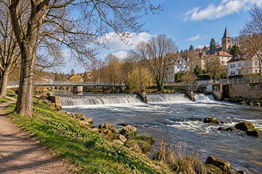 A river with a waterfall, a bridge, trees, and buildings in the background. 2